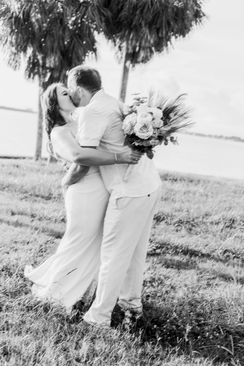 Black and white wedding photo showing couple embracing in a romantic moment with palm trees and floral bouquet.