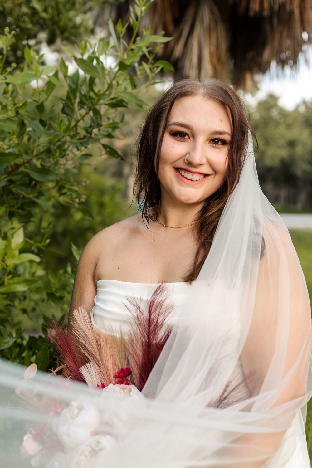 A bride smiles outdoors in a white strapless gown with a flowing veil and burgundy feather bouquet details.