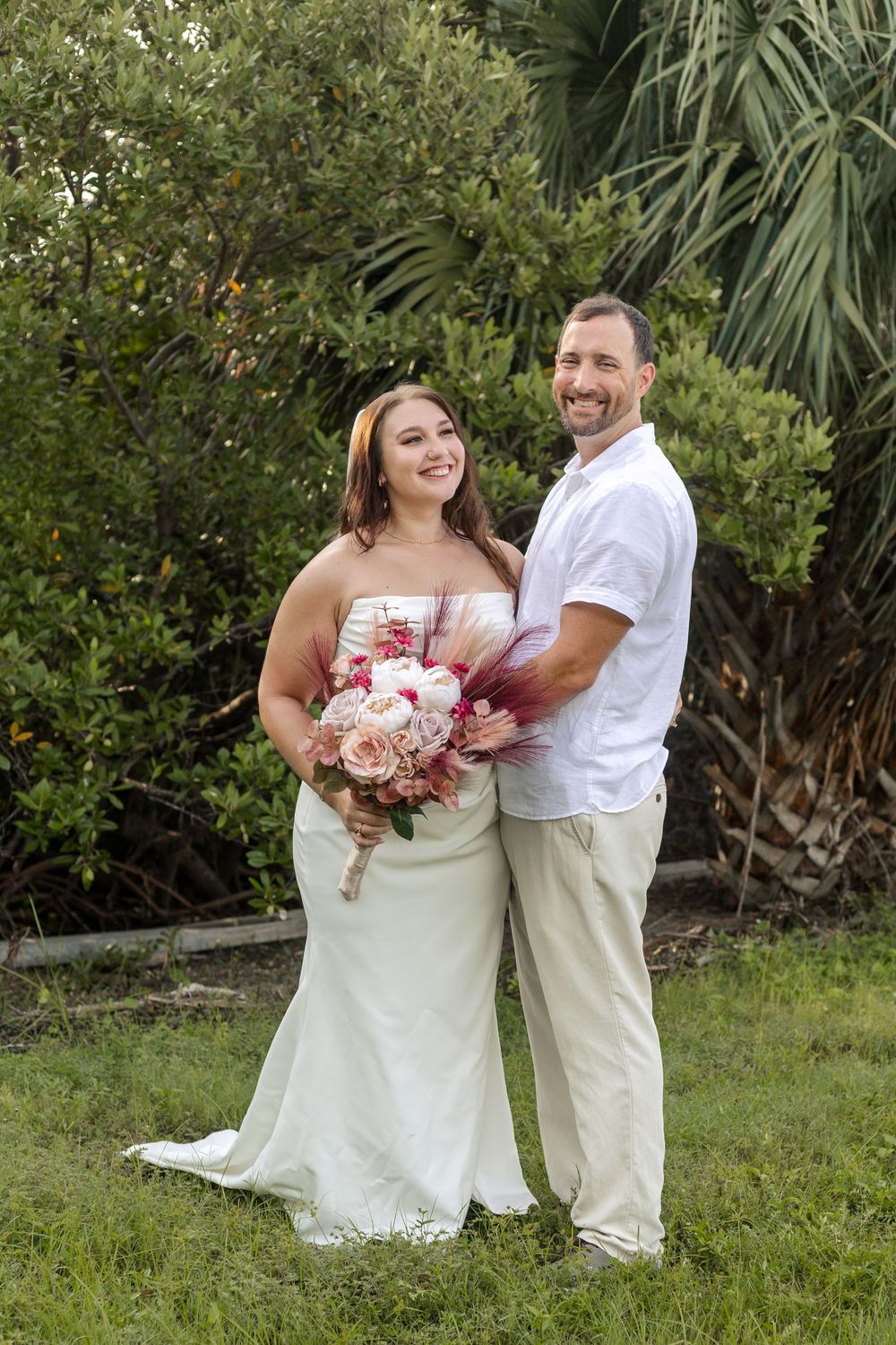 A couple poses outdoors for a wedding photo with palm trees and greenery in the background.