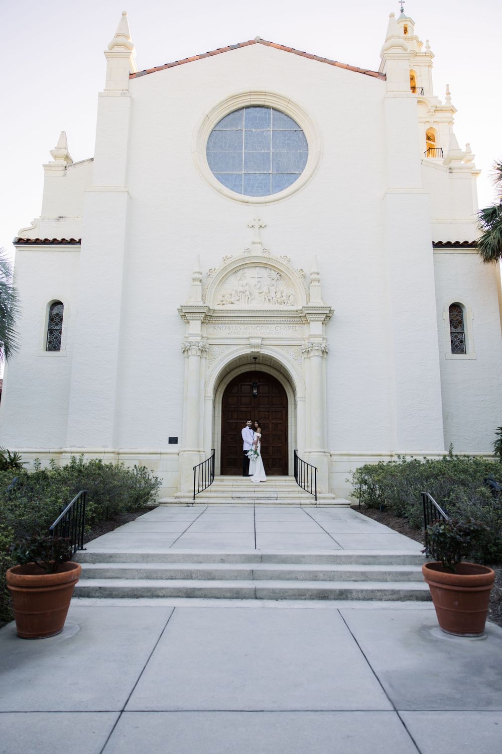 Spanish colonial church with white facade, round window, and two figures standing in the elegant arched doorway entrance.