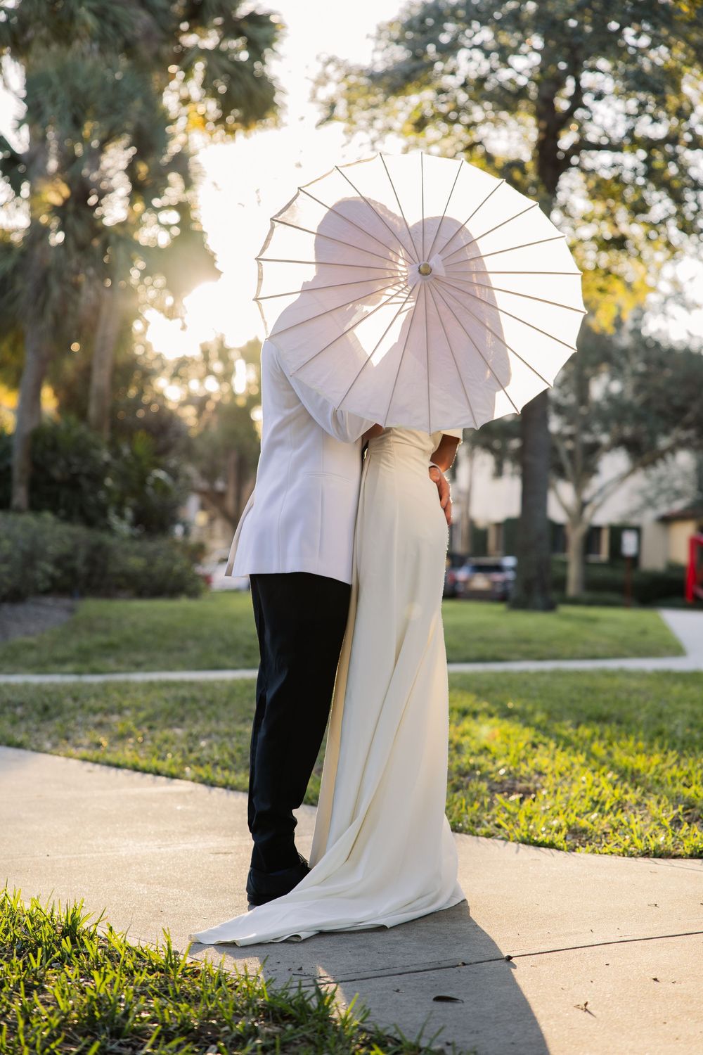 A romantic couple embraces under a white parasol on a sidewalk during golden hour with trees in the background.
