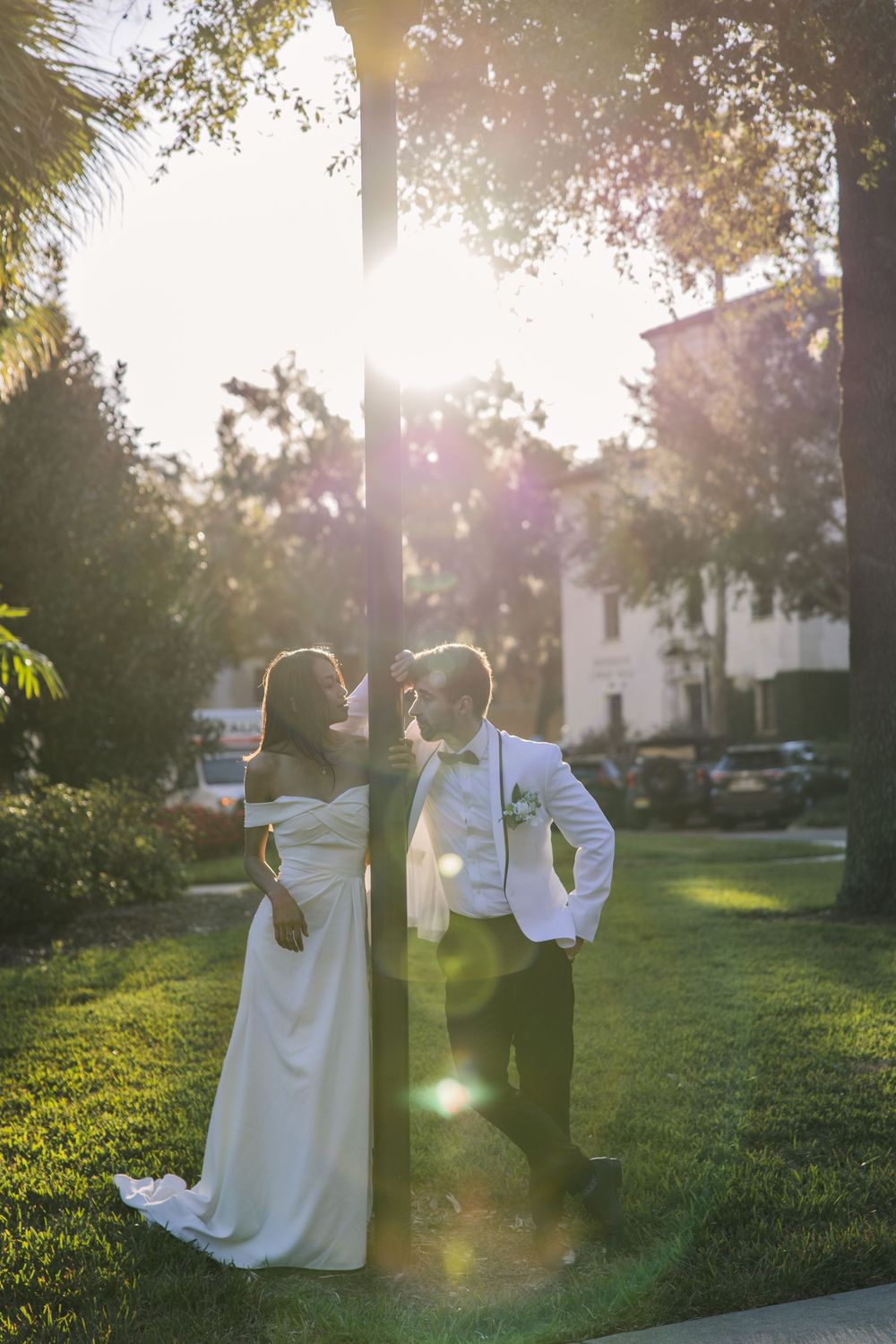 A romantic sunset wedding photo of a couple in wedding attire standing on a lush green lawn with sunlight streaming through trees.