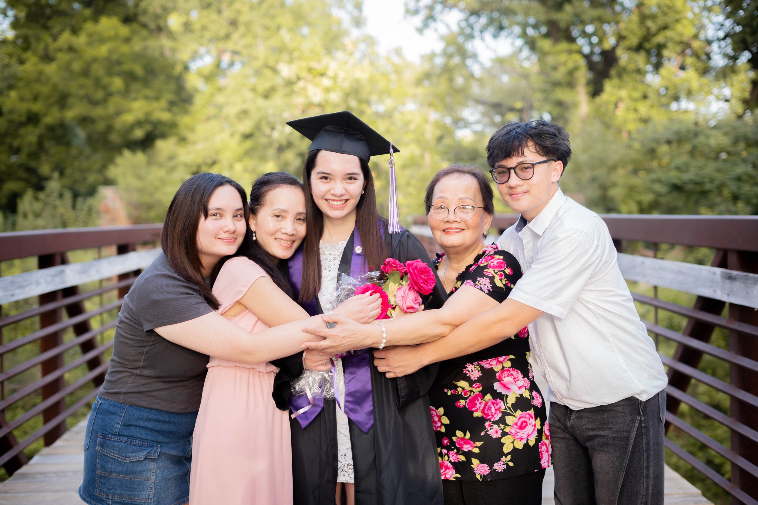 Happy graduate in cap and gown celebrating with four family members on a wooden bridge outdoors.