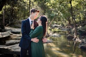 Romantic couple nearly kissing outdoors near a rocky creek, woman in green dress and man in navy suit.