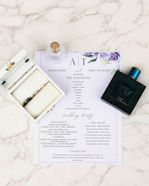 Wedding invitation suite with purple floral details and perfume bottle on marble background.