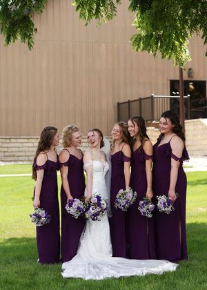 Bridesmaids in deep purple dresses share a candid laughing moment with the bride outdoors.