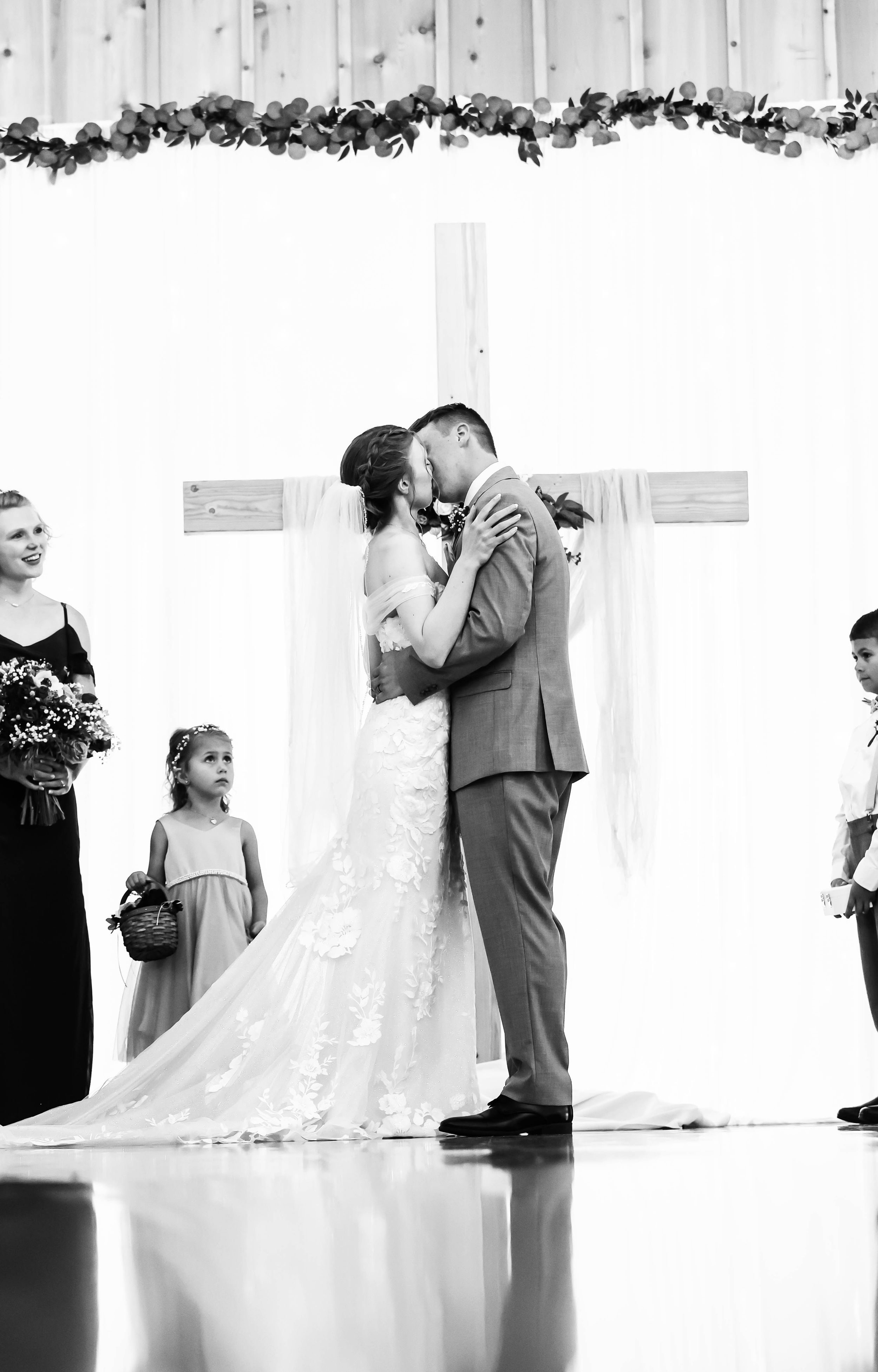 Black and white ceremony photo with couple kissing under cross and floral arch.