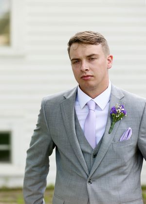 Groom in grey suit with purple boutonniere and pocket square against white building.