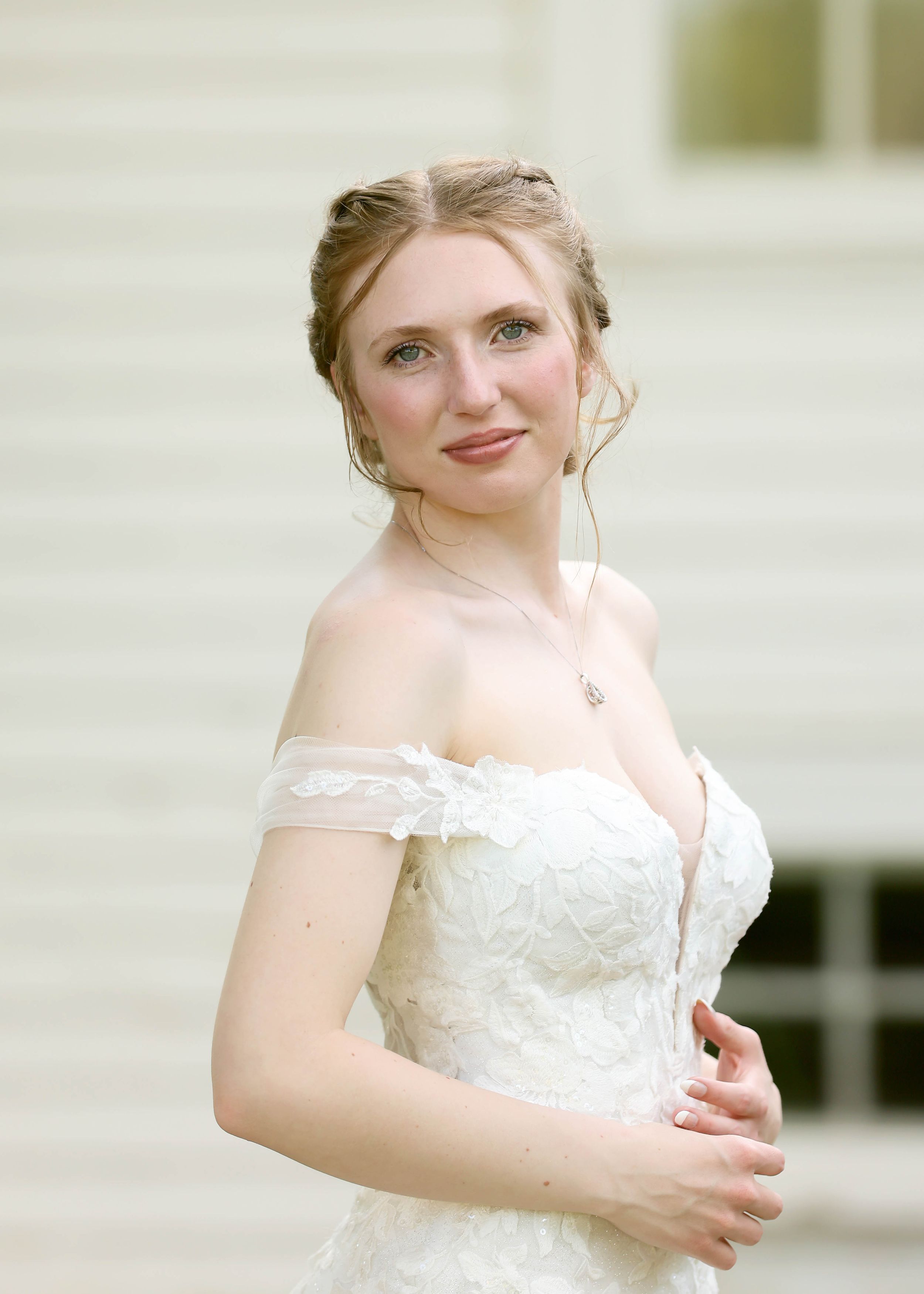 Bride in off-shoulder white lace wedding gown posing against white building backdrop.