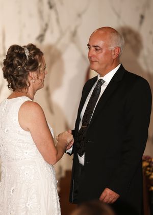 Couple exchanging vows at wedding ceremony, groom in black suit with tie holding brides hands, bride in white lace gown.
