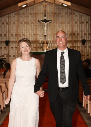 Smiling couple holding hands walking down church aisle, bride in white gown and groom in black suit with tie.
