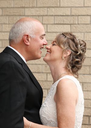 A joyful couple in wedding attire shares a romantic moment, smiling at each other against a brick wall backdrop.