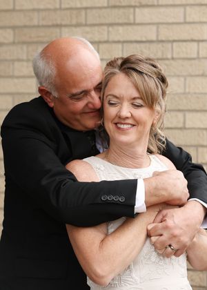 A couple embraces tenderly in wedding attire, the groom nuzzling the smiling bride against a brick wall backdrop.