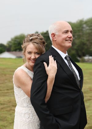 A smiling bride in white lace gown stands behind a groom in black suit outdoors on their wedding day.