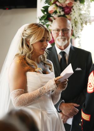 A bride in a lace dress reads from a paper during an indoor wedding ceremony.
