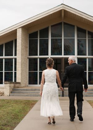 Bride in white dress and groom in black suit walk hand-in-hand toward a modern church on their wedding day.