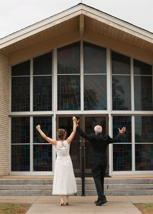 Newlywed couple in wedding attire raising hands in celebration outside a church with stained glass windows.