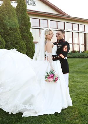 A flowing white wedding dress billows in the wind on a grassy lawn with a building in the background.