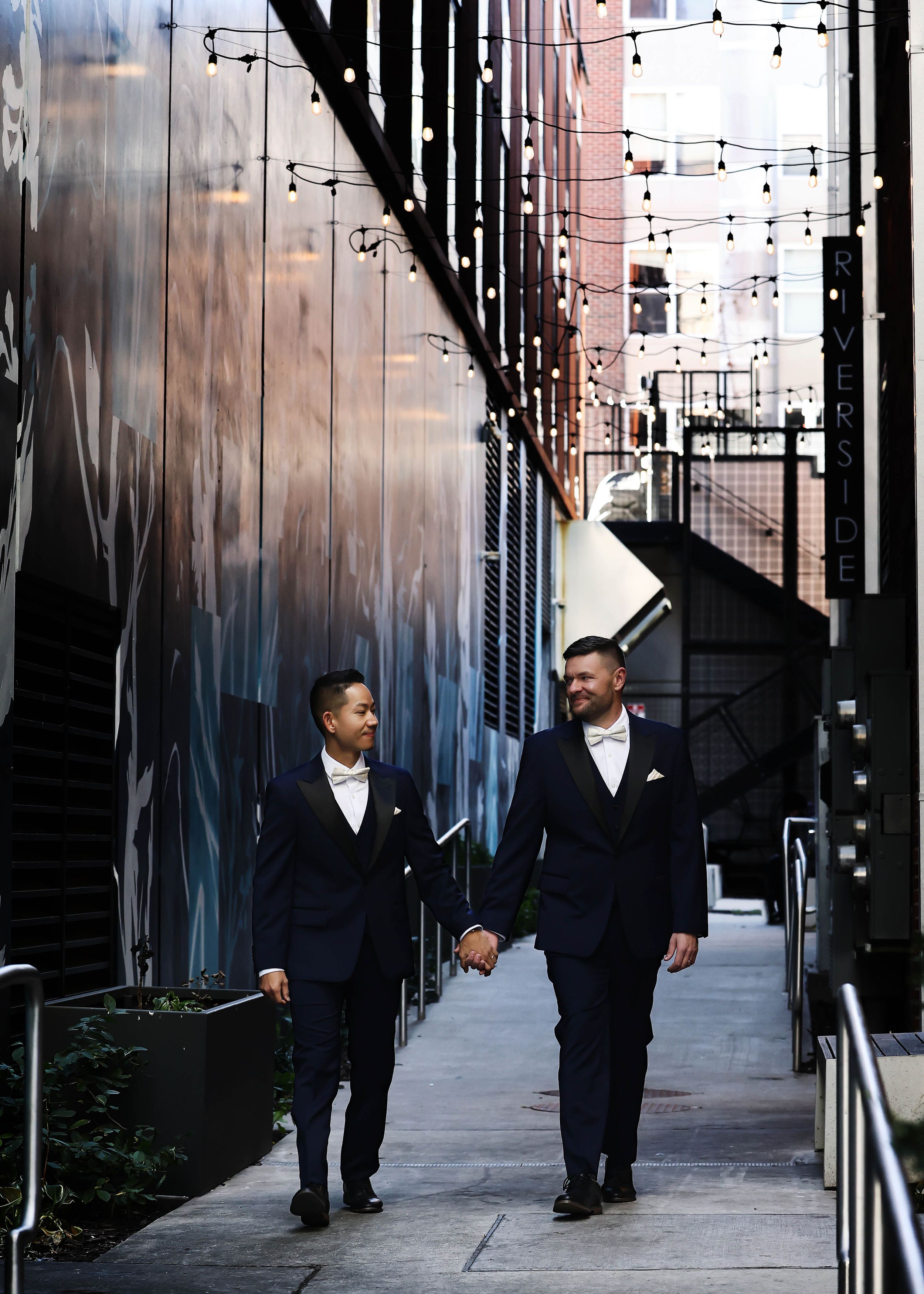 Couple in formal wear walking along industrial pathway with string lights overhead.