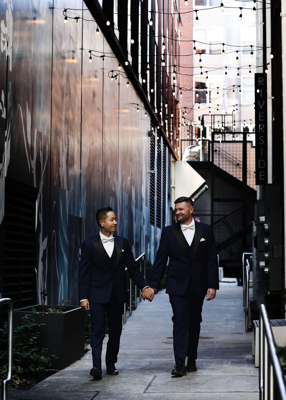 Couple in formal wear walking along industrial pathway with string lights overhead.