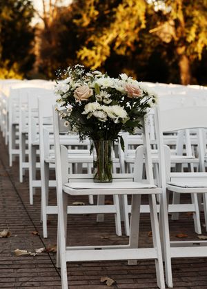 White folding chairs line an outdoor ceremony aisle with a floral arrangement in a glass vase.