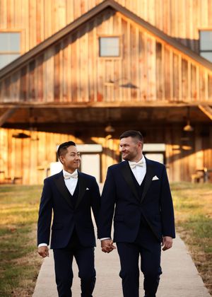 Two people in matching navy suits walk hand in hand on a path leading to a rustic wooden barn.