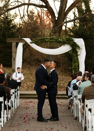 Wedding ceremony under a white draped arch with greenery in a wooded setting.