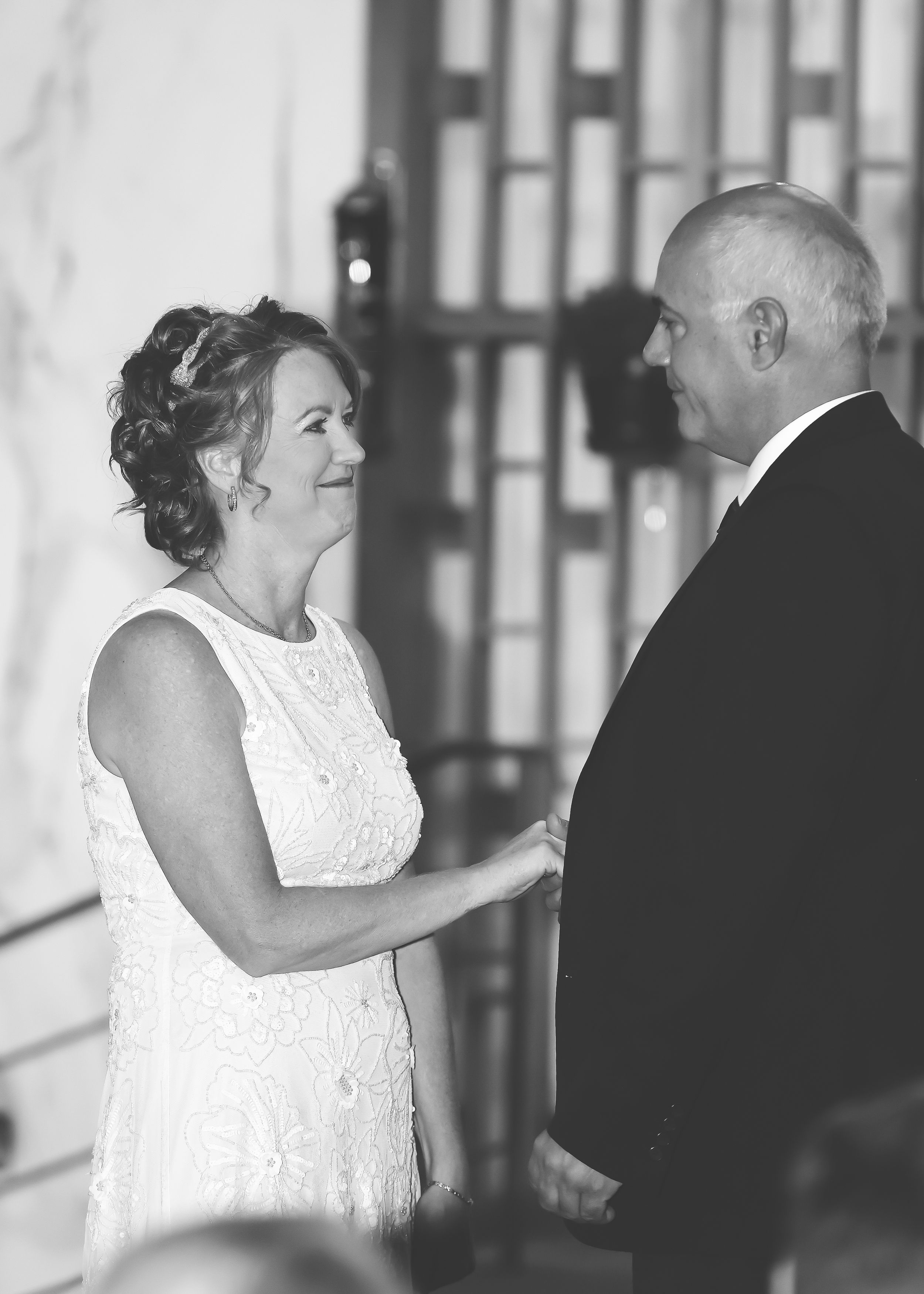 Black and white photo of a smiling bride in white lace dress exchanging vows with bald groom at wedding ceremony.