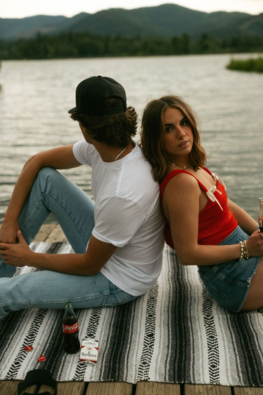 Two people sitting back to back on a blanket by a lake with mountains in the background at dusk.