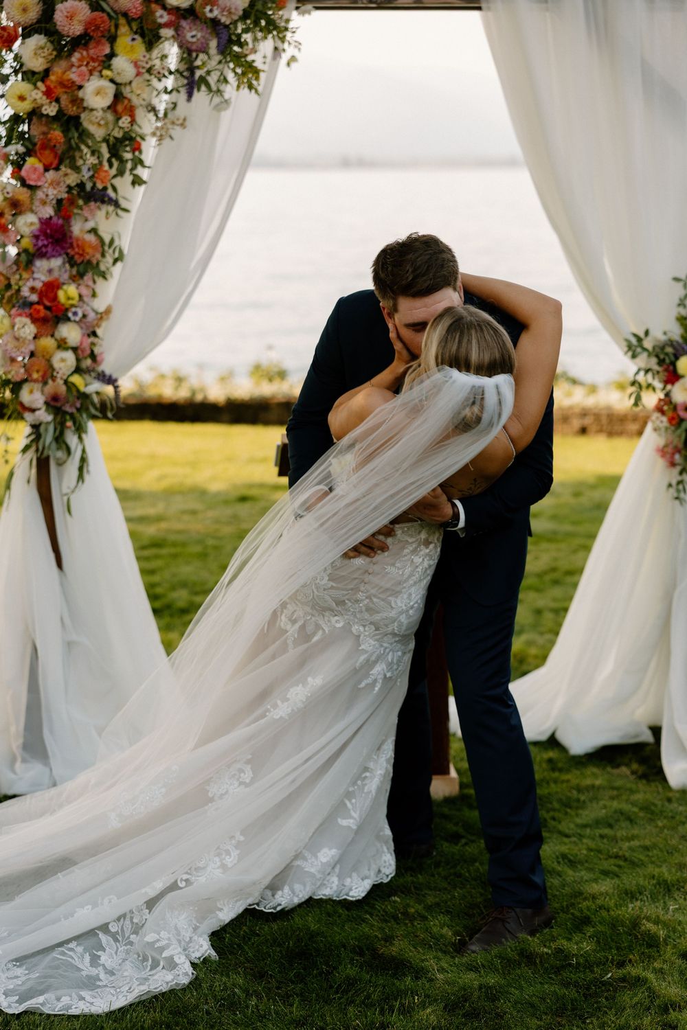 A romantic wedding kiss sequence showing the couple embracing under white floral arches at an outdoor ceremony.