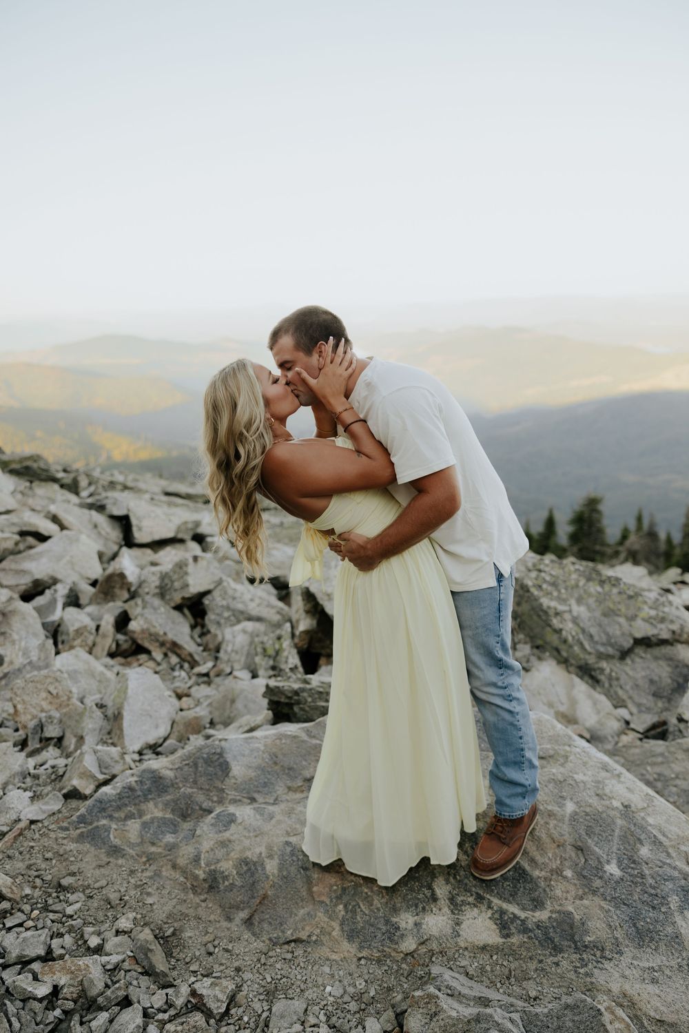 A couple shares an intimate moment on rocky mountain terrain at sunset with scenic mountain views in the background.
