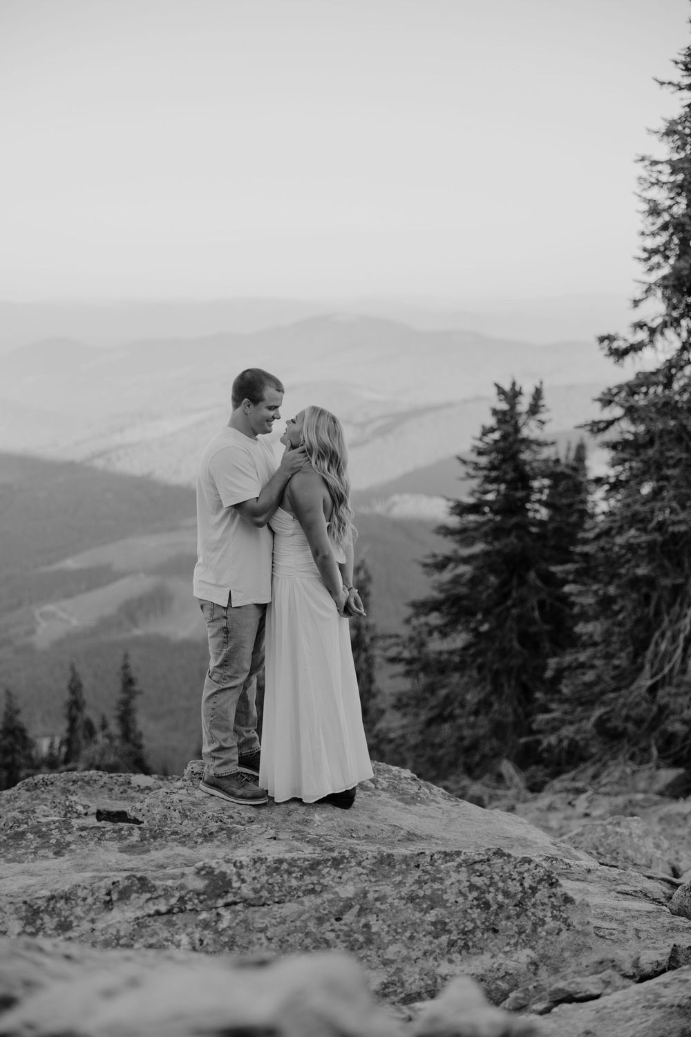 A romantic black and white mountain vista shows a couple in light clothing embracing on a rocky overlook surrounded by evergreens.