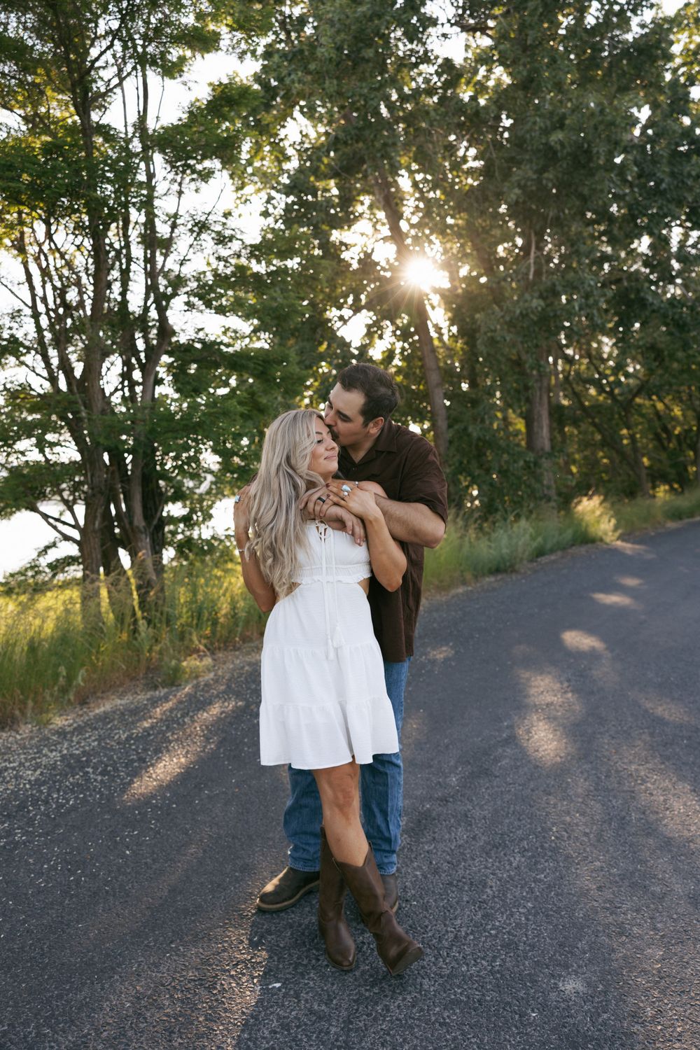 A couple embraces on a sunset-lit country road surrounded by green trees.