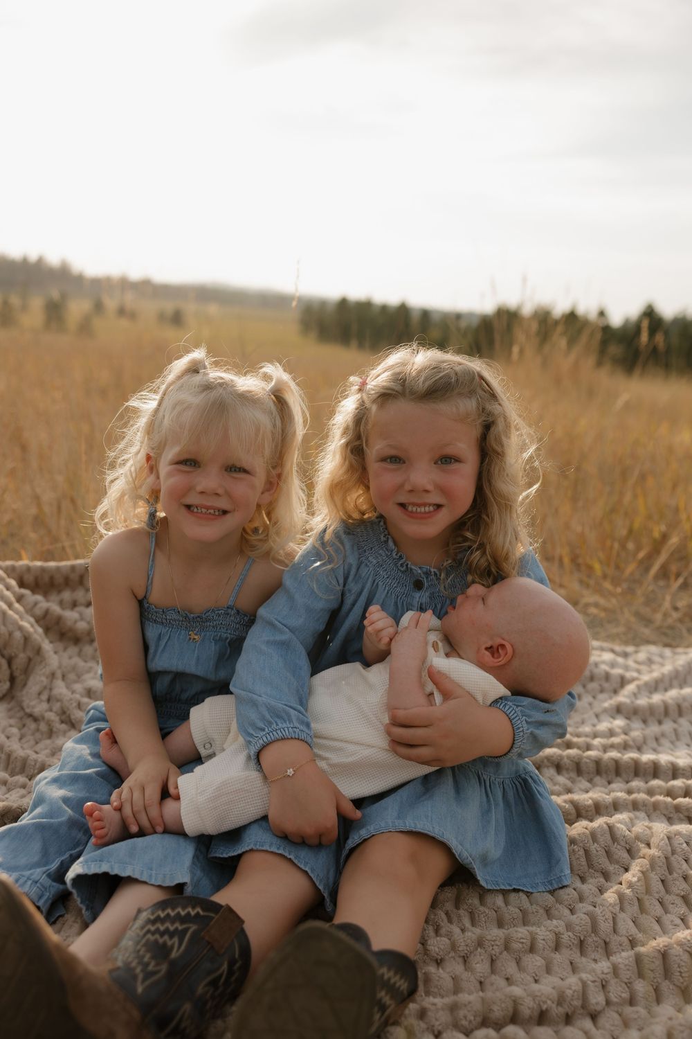 Small children in denim dresses sitting together on a blanket in a golden field at sunset.