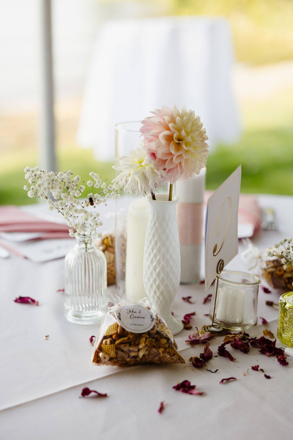Elegant wedding table setting with white vases, pink dahlia flower, and scattered rose petals on white tablecloth.