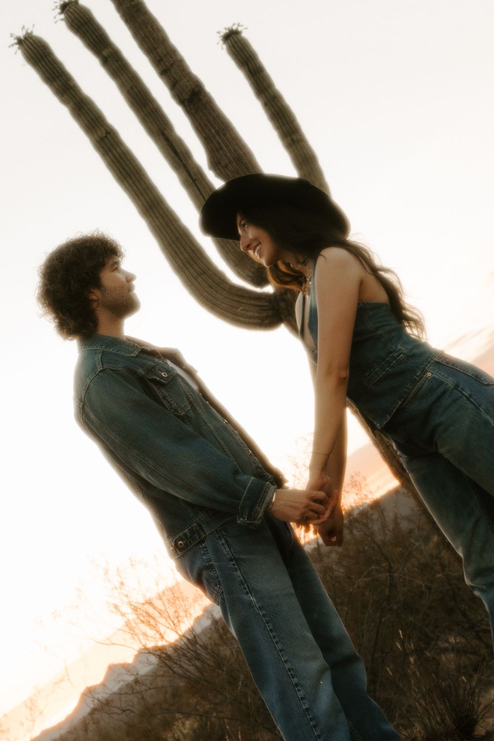 Two people in winter clothing lean back against each other with arms interlocked against a bright sunset sky.