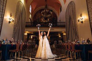 A couple stands in a grand ballroom with elegant arched windows, ornate chandeliers and rows of chairs for a wedding ceremony.