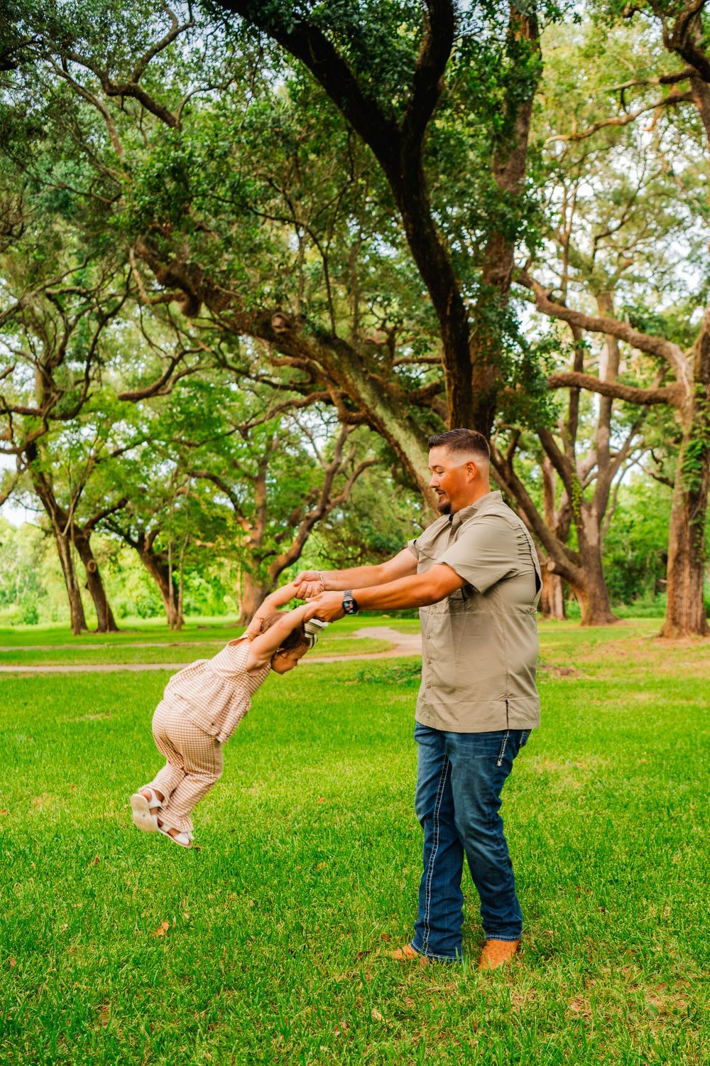 A father joyfully swings his young child in a lush green park surrounded by large oak trees.