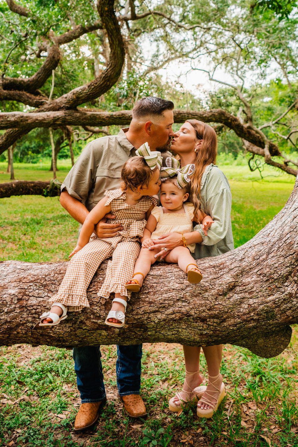 A couple kisses while sitting on a large tree branch in a lush park, each holding a young daughter in matching outfits.