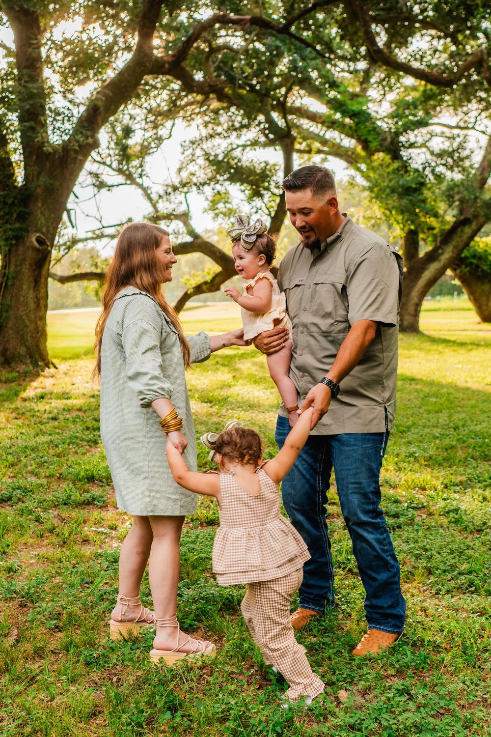 Family of four enjoying a sunny outdoor photo session under large oak trees in a lush green park.
