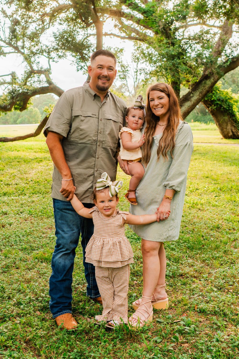 Family of four posing outdoors under large oak trees on green grass, parents with two young toddler girls in coordinated outfits.