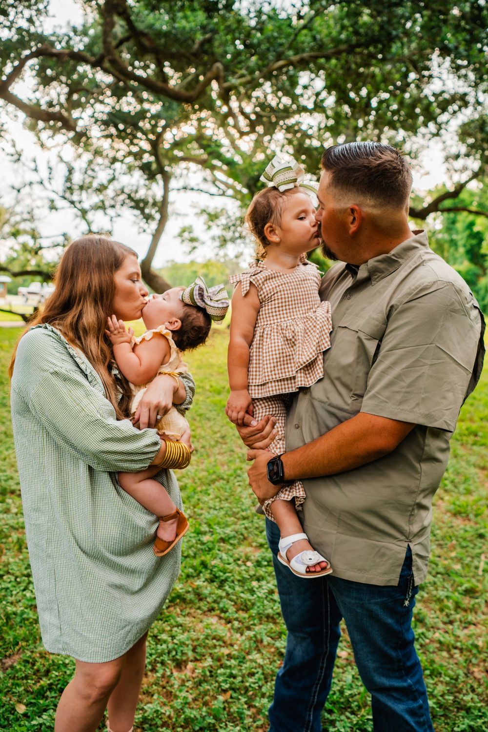 A young family of four poses outdoors, parents holding two small children who share a sweet kiss under a large oak tree.
