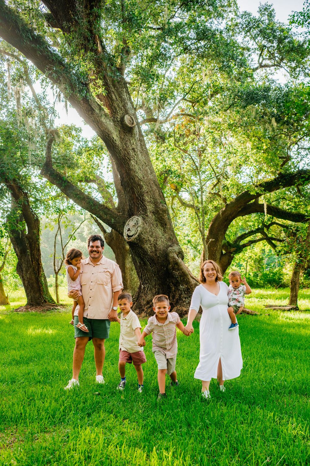 Family of six walks on lush green grass beneath sprawling oak trees, parents holding children's hands outdoors.