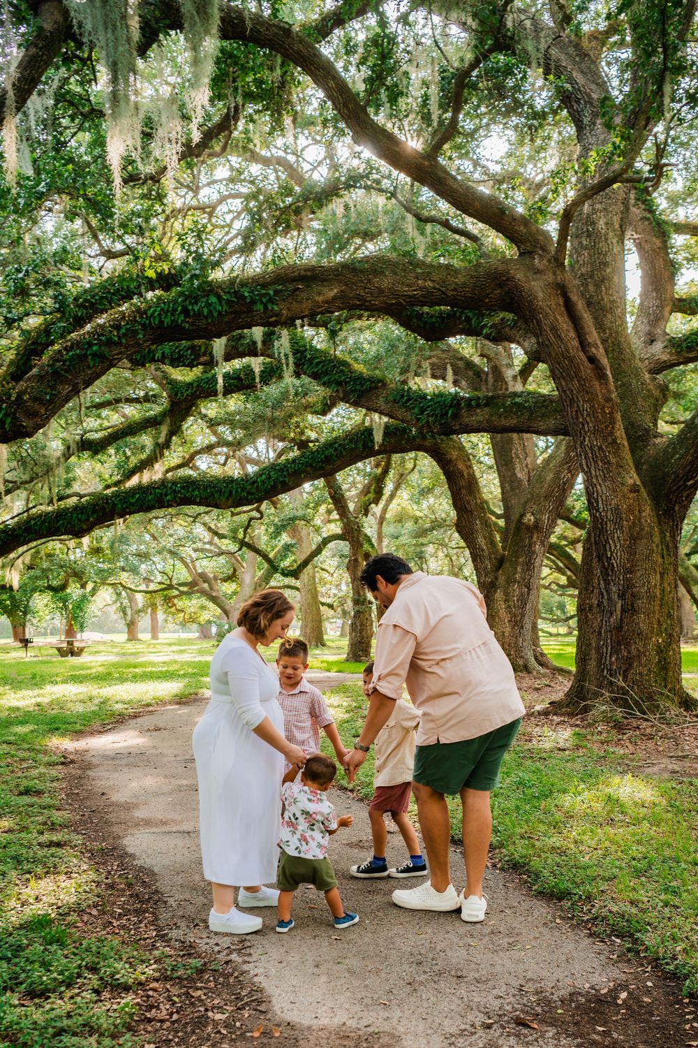 Family with two young children walking on a path under sprawling moss-covered oak trees in a lush green park.
