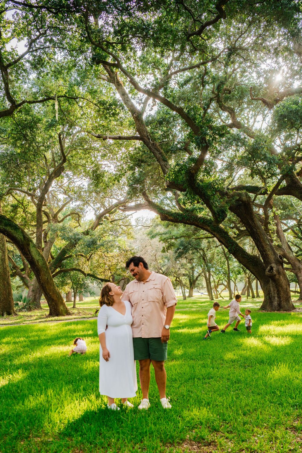 Couple stands under sprawling oak trees on lush green grass in a sunlit park, with children playing in the background.