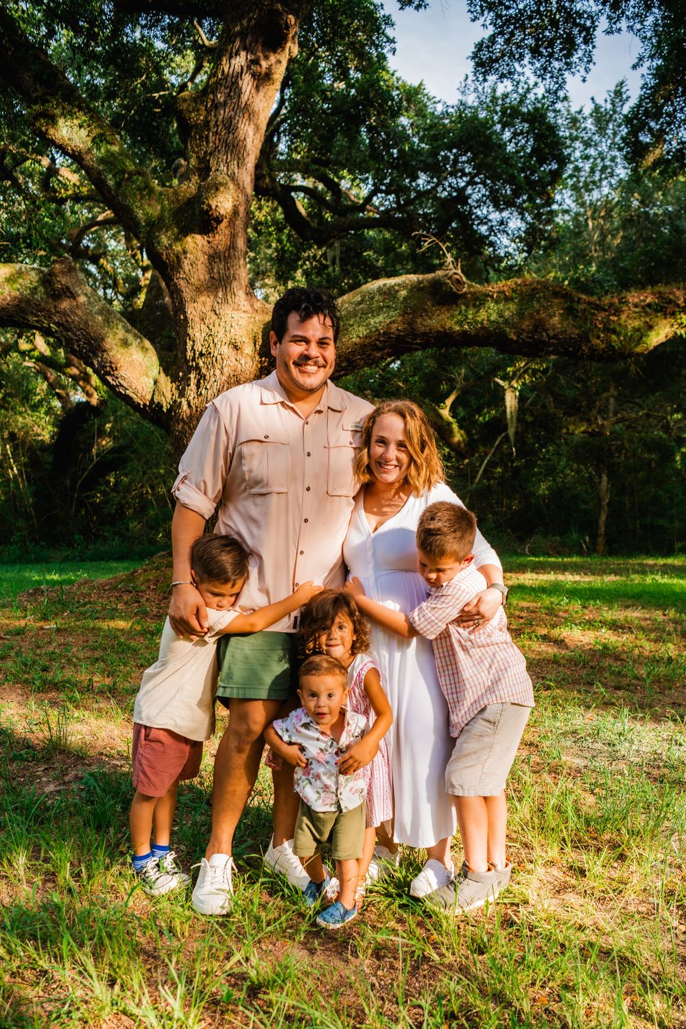 Happy family of five posing outdoors under a large oak tree on a sunny day in a lush green park.