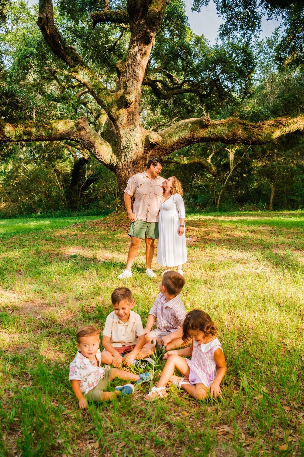 Family of five poses under a large oak tree in a sunny green park, parents kissing while three children play on the grass.