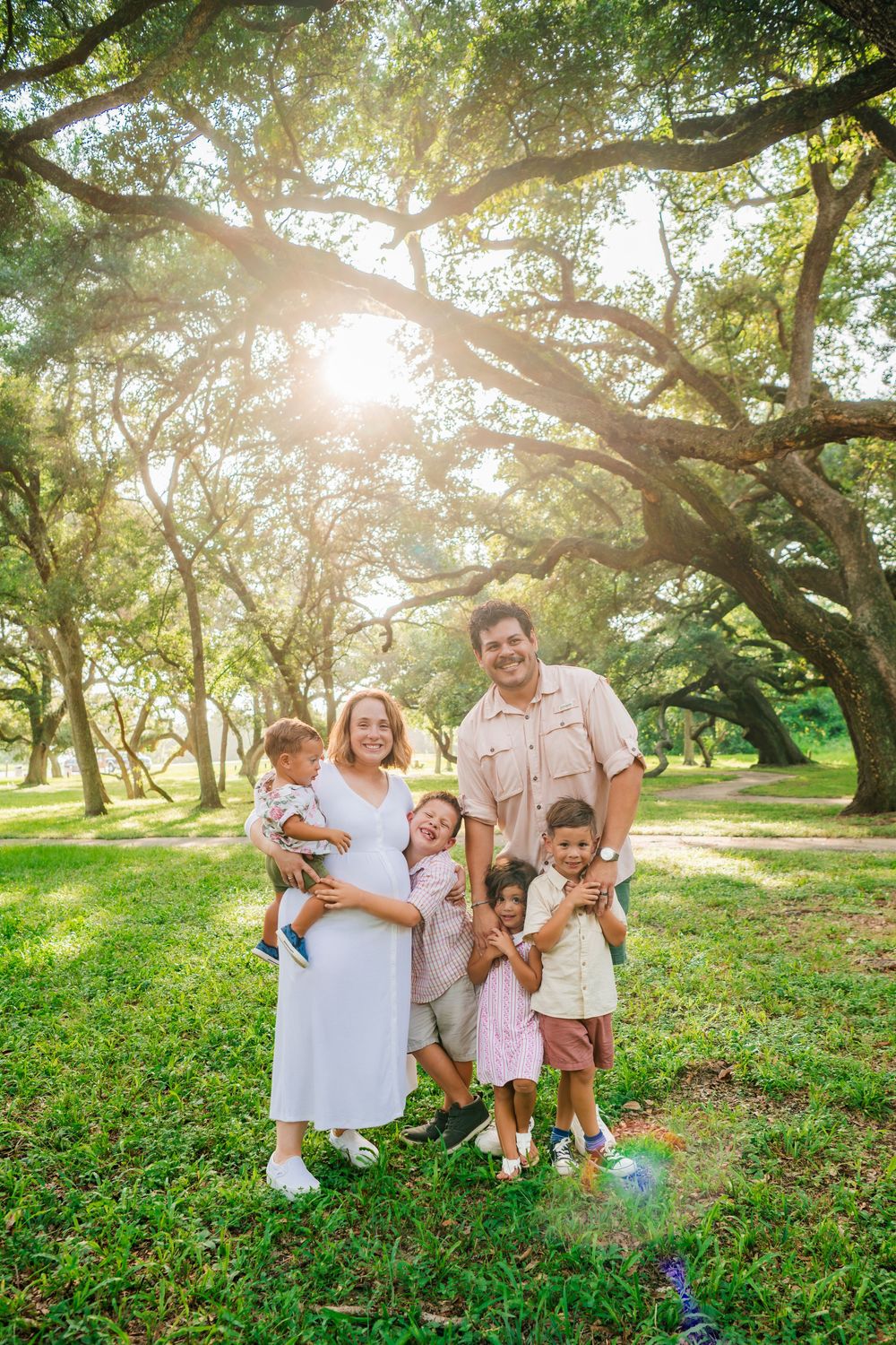 Happy family of six posing outdoors under large oak trees with sunlight streaming through the canopy on green grass.