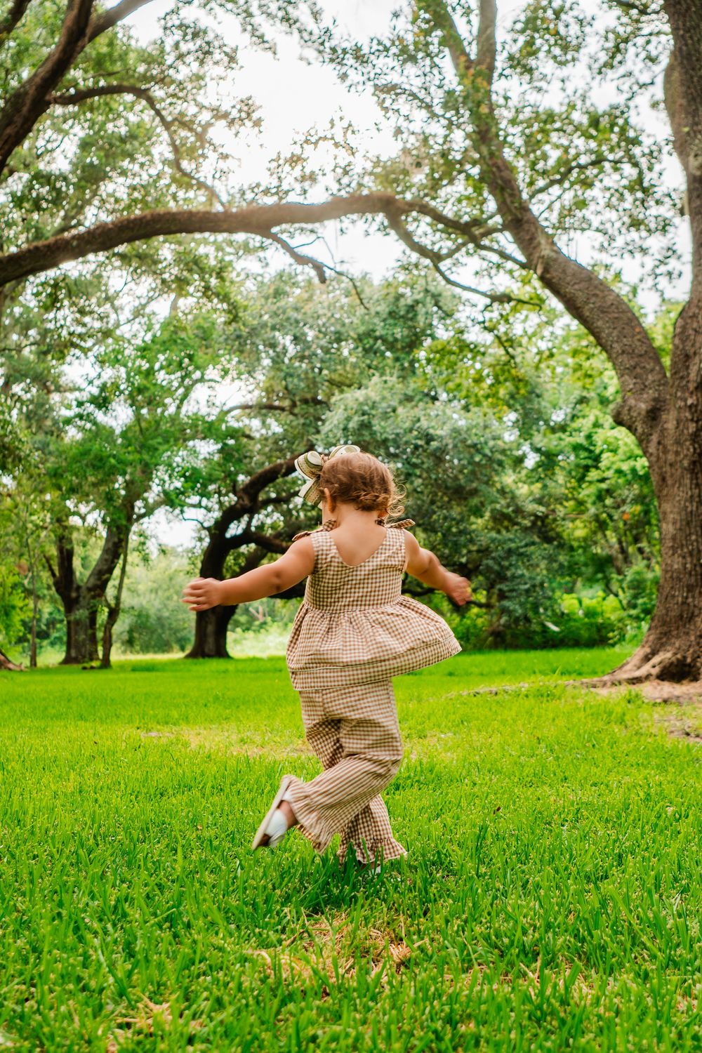A young girl in a plaid romper runs joyfully across lush green grass beneath large oak trees in a sunny park.