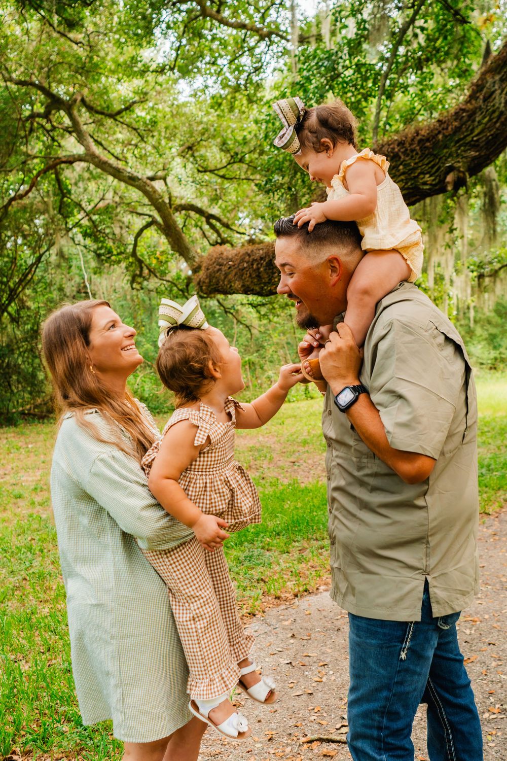 A happy family of four poses outdoors, with parents holding two young girls amid lush green trees.